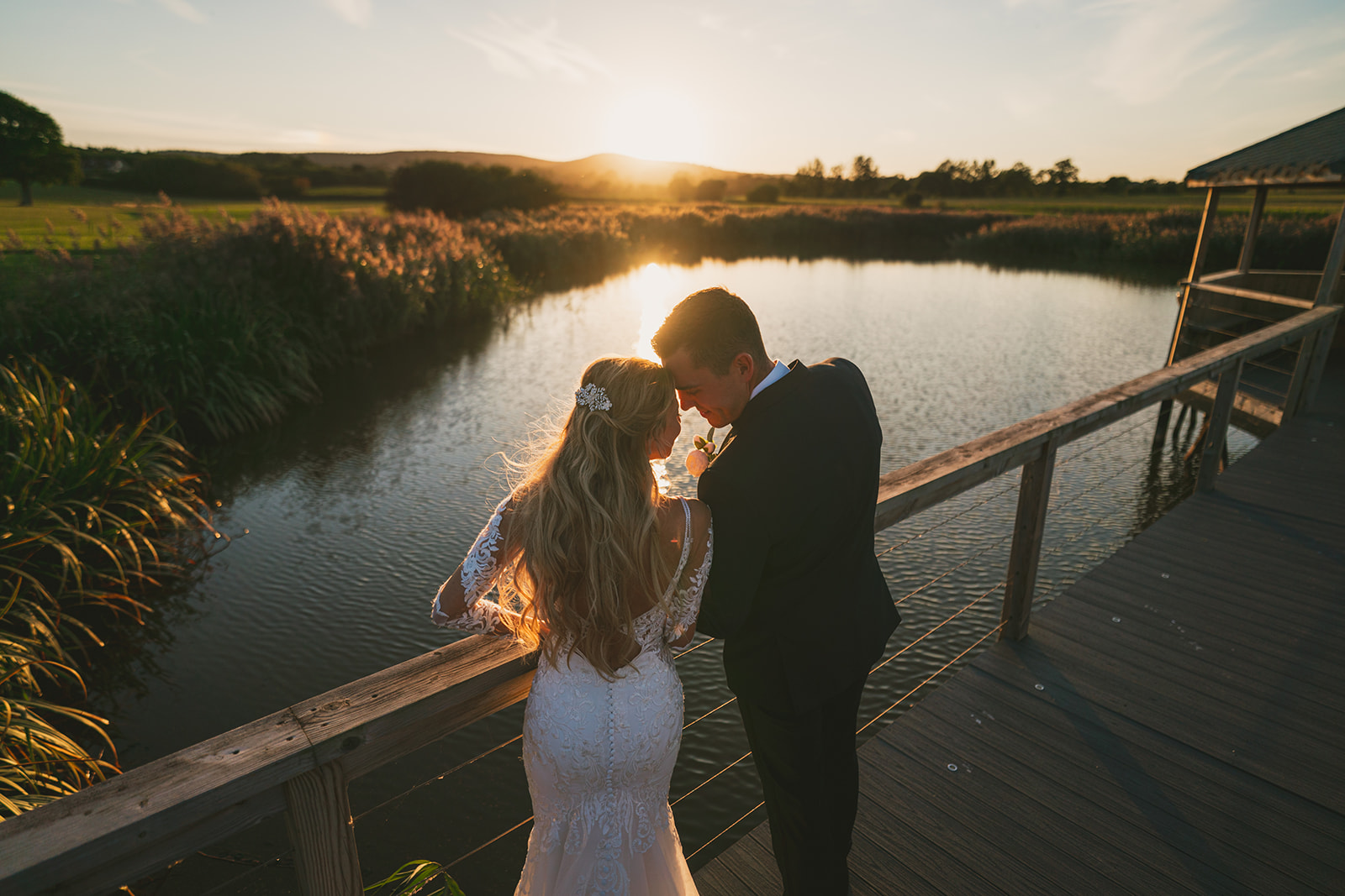 A bride and groom standing on a bridge overlooking a pond at sunset.