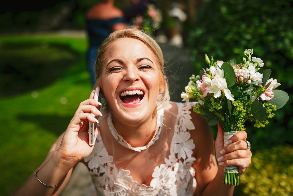 A bride laughing while holding a bouquet and talking on the phone, captured by a natural wedding photographer in Wiltshire.