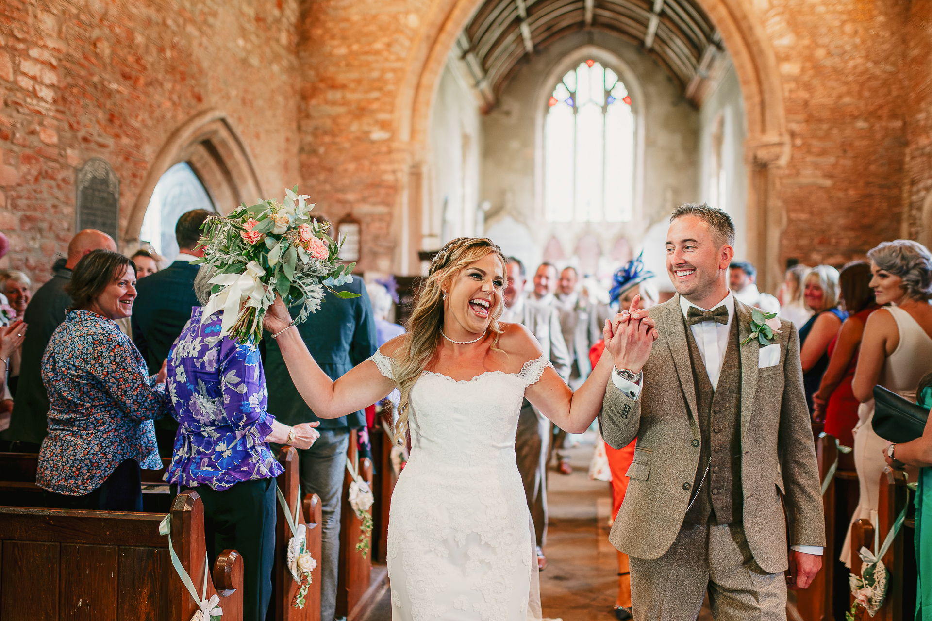 A bride celebrate by punching the air with her flowers as she walks down the aisle of a church after just getting married in a church near Bristol