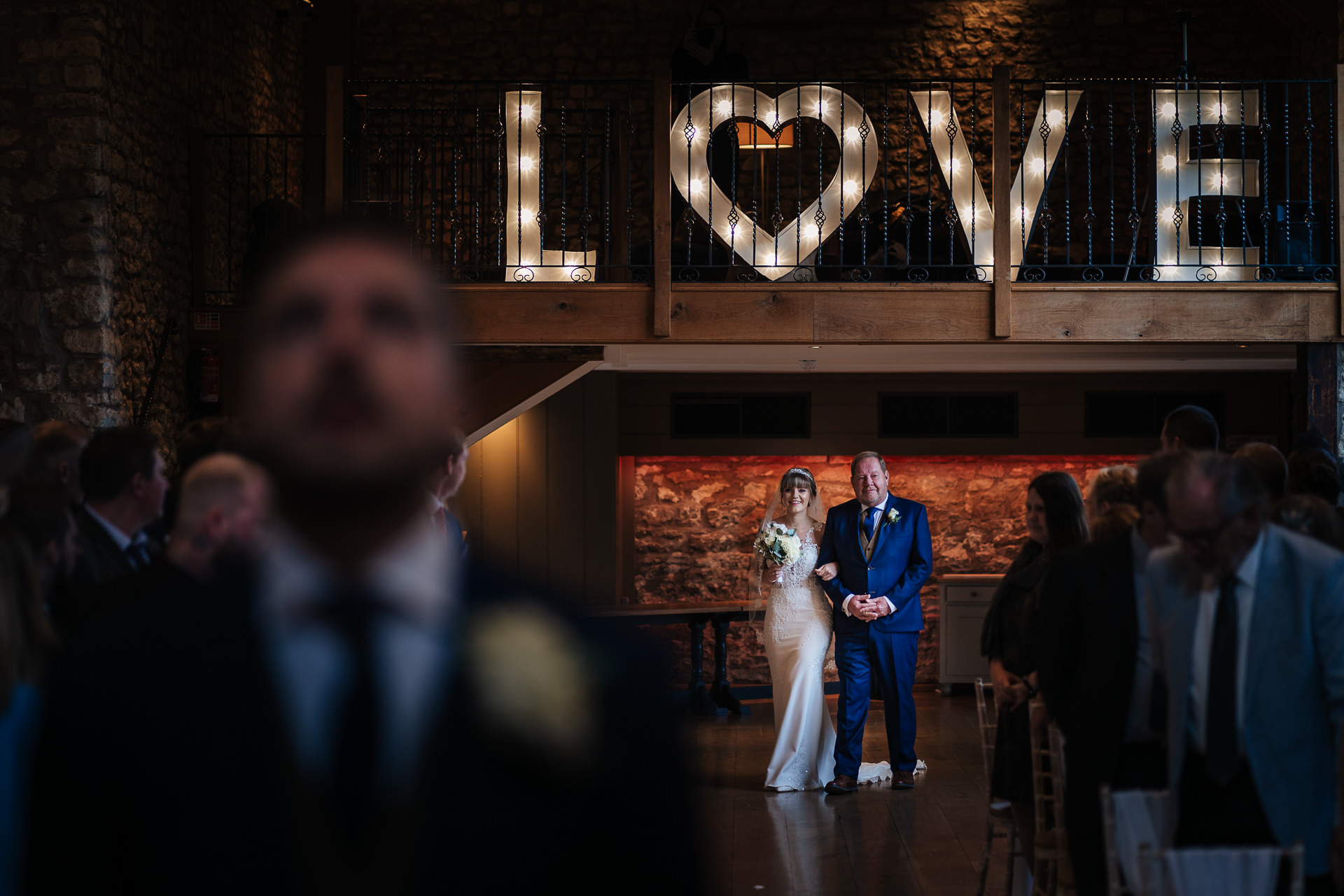 A bride and groom walking down the aisle in front of a lit up love sign.