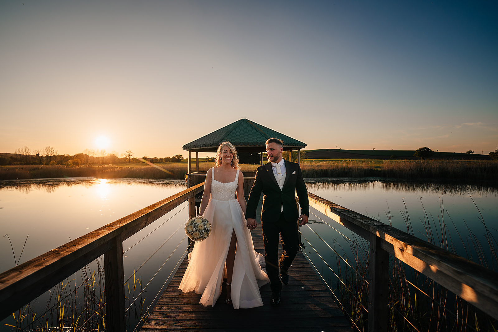 A bride and groom hold hands walking down a wooden pier at sunset, with a gazebo and still water in the background. The bride holds a bouquet and both are smiling.