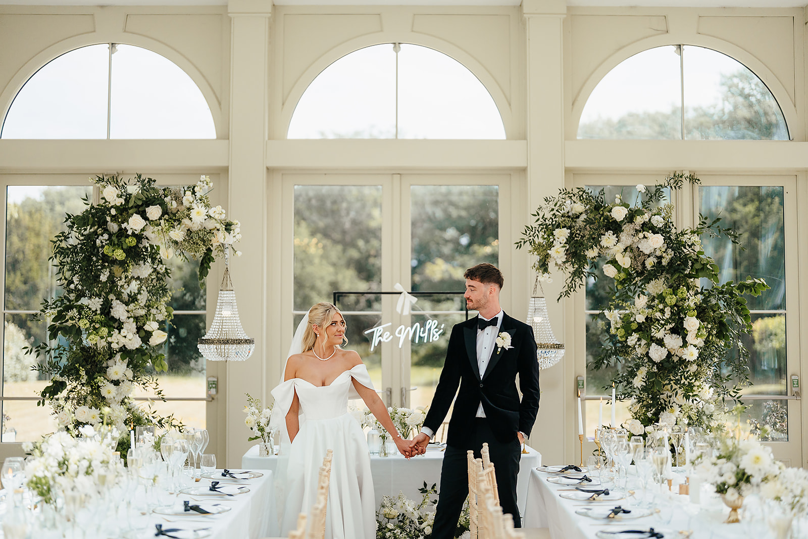 A bride and groom stand holding hands at a decorated wedding reception table, with floral arrangements and a "Be Mine" sign behind them in a bright, windowed room