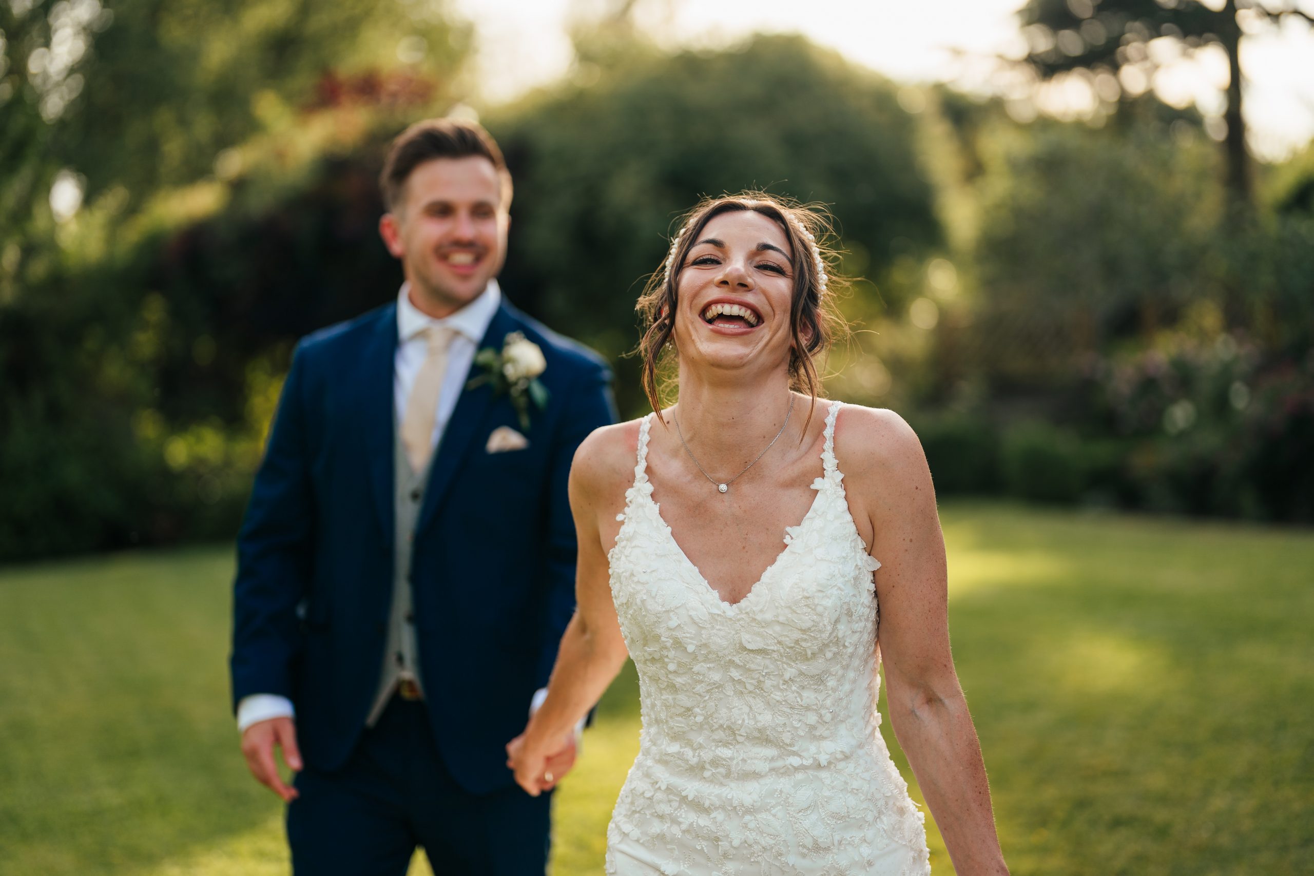 A woman in a white wedding dress smiles and laughs while holding hands with a man in a blue suit outdoors in a garden setting