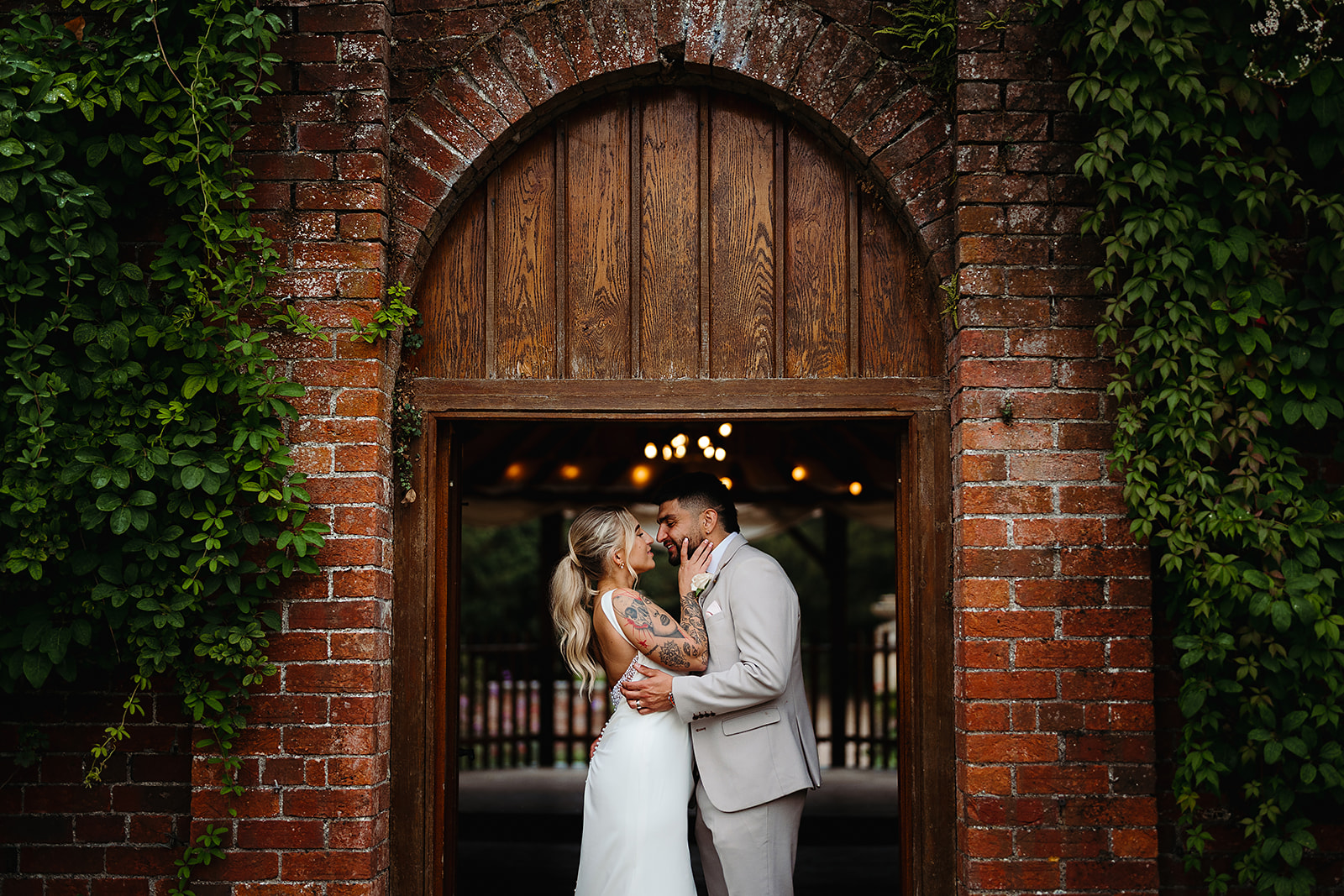 A bride and groom stand closely together, embracing and smiling under a wooden archway set in a brick wall with green vines