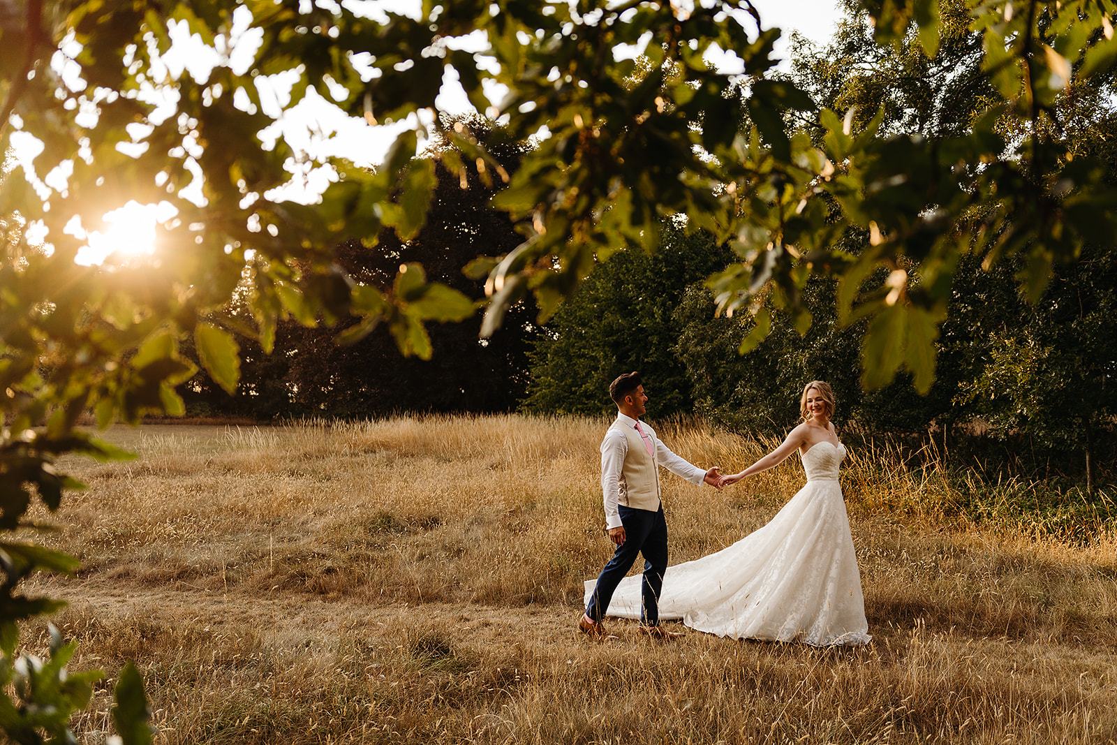 A bride and groom walk hand in hand through a sunlit grassy field, framed by tree branches in the foreground