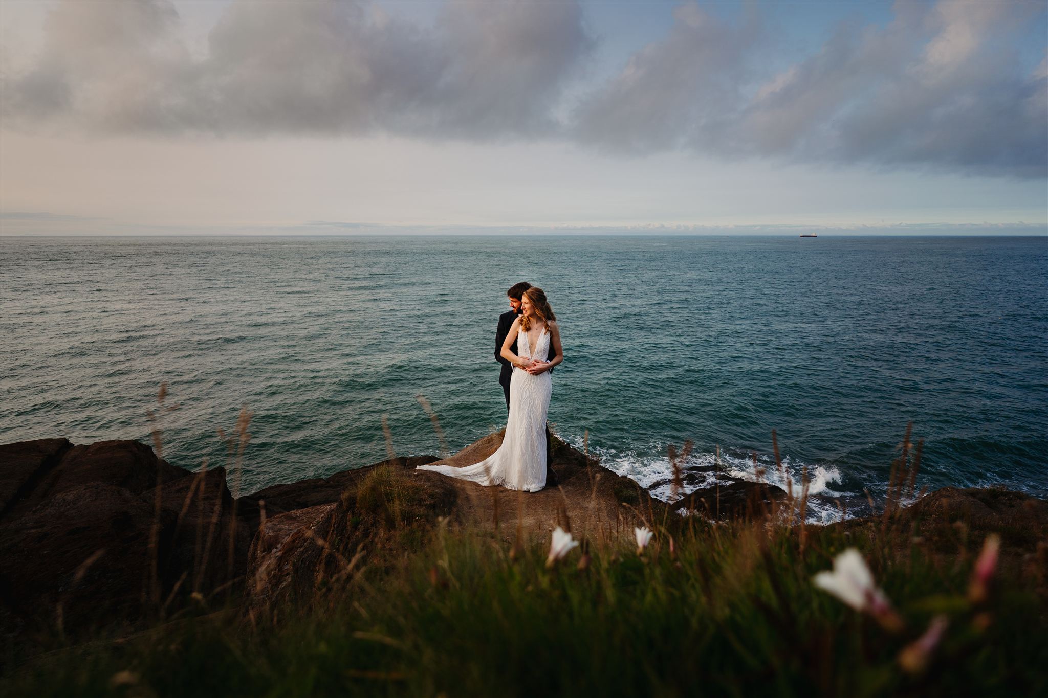 A couple in formal wedding attire stands on rocky cliffs by the ocean, with waves and a cloudy sky in the background