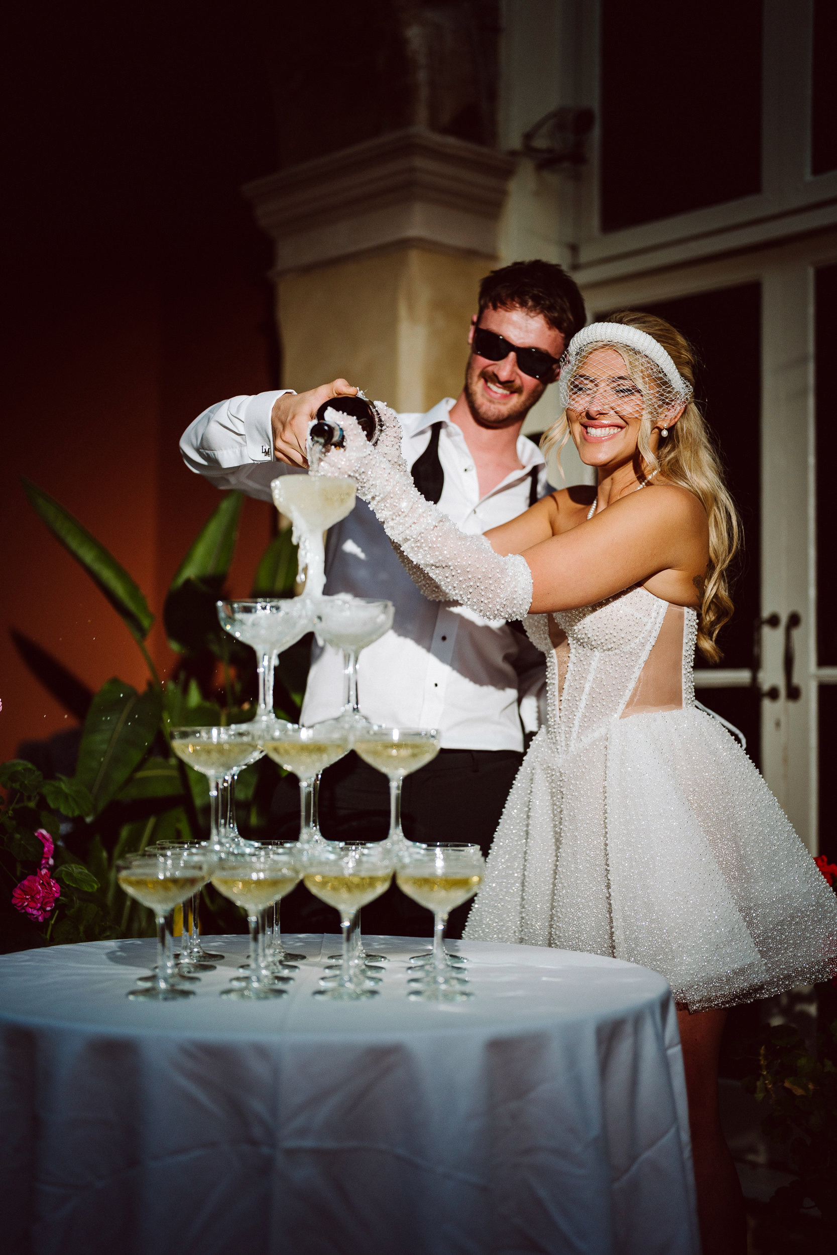 A man and woman dressed in formal attire pour champagne into a tower of glasses at an outdoor event, smiling at the camera