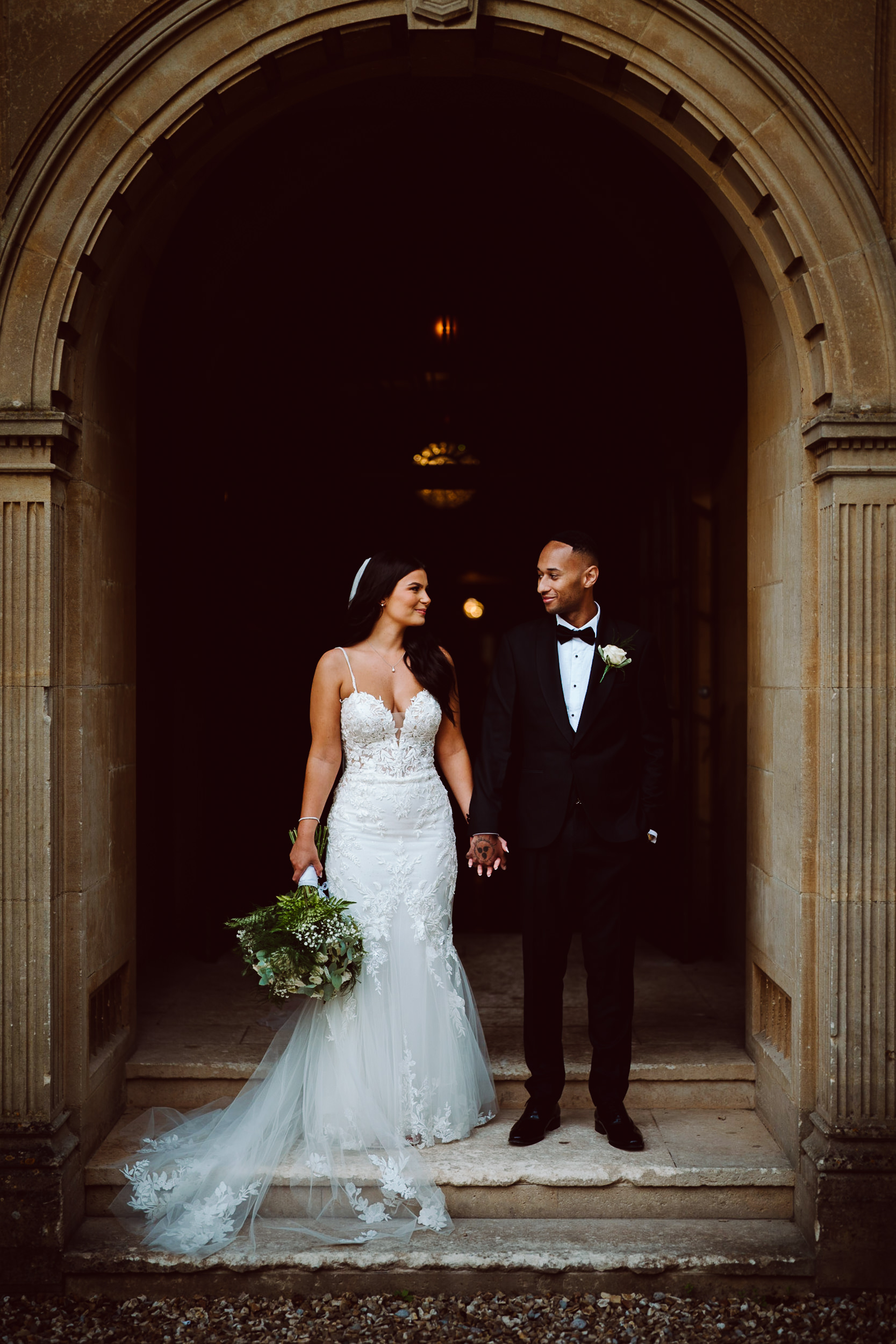 A bride and groom stand holding hands under a stone archway; the bride wears a white dress and holds a bouquet, and the groom wears a black tuxedo