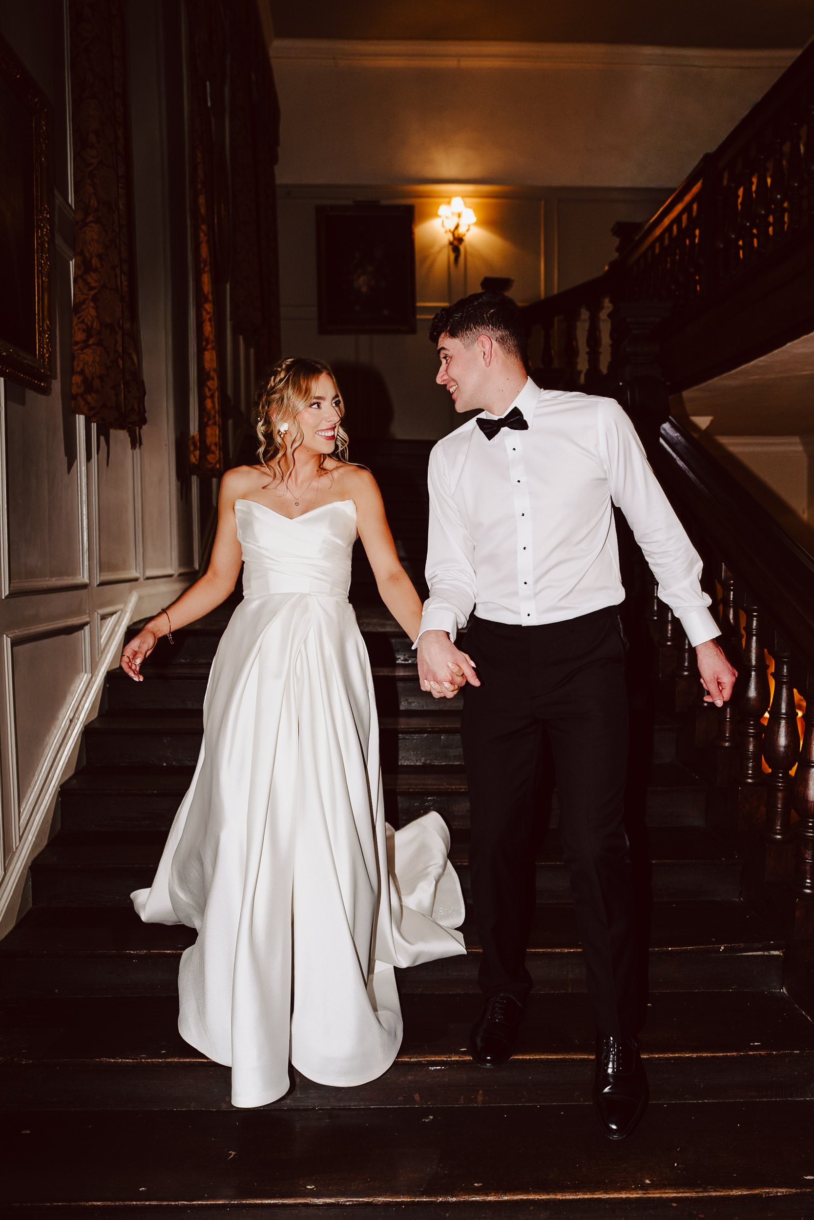 A bride in a white dress and a groom in a white shirt and black trousers walk hand in hand down a staircase in an elegant indoor setting