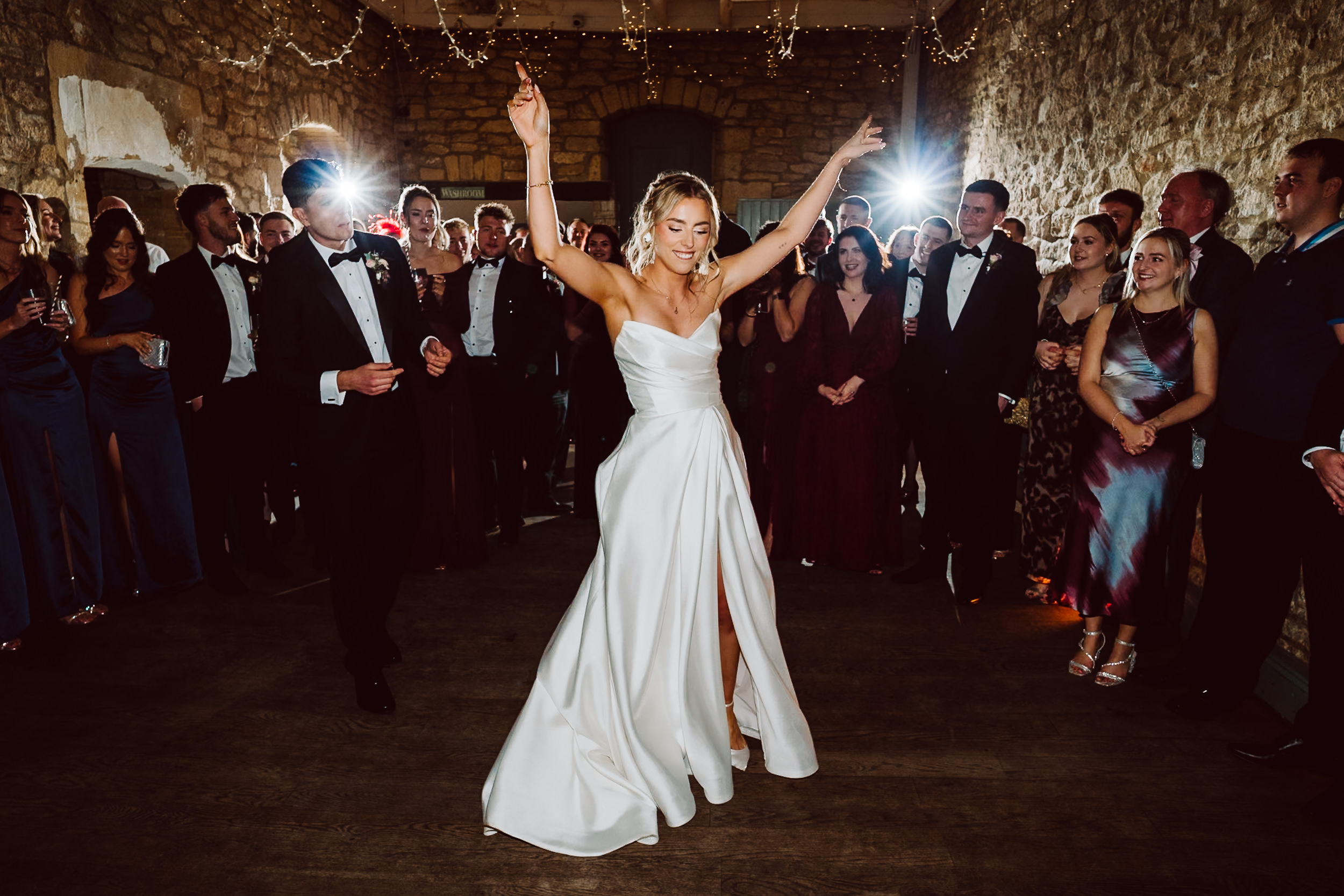 A bride in a white strapless dress dances with her arms raised while wedding guests in formal attire watch and smile in a stone walled venue