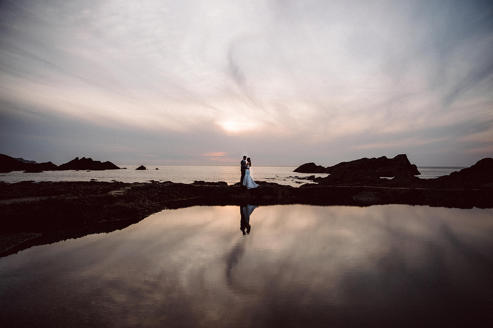A couple stand together by the water at sunset, with their reflection visible in a still pool and rocky coastline in the background